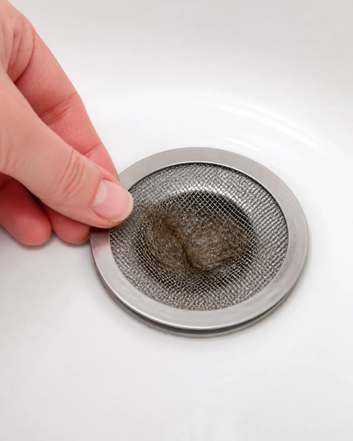 a close-up of a stainless steel mesh drain cover sitting in a white bathtub drain with a clump of wet dog fur collected on top of the mesh clearly showing how it catches hair before it enters the pipe and a persons hand about to lift the mesh to empty it bright overhead bathroom light